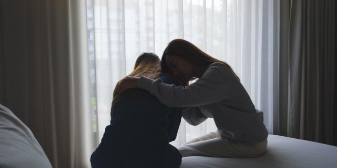 Silhouettes of two people in a dark room sitting on the edge of a bed in front of a bright window hugging each other while experiencing the effects of the most addictive painkillers