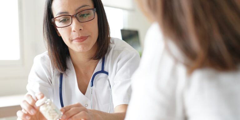 Female doctor wearing glasses and a stethoscope discusses medication with a patient, holding a pill bottle while explaining dosage or treatment details.